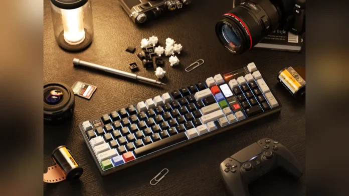 Lifestyle desktop setup showing QK100 Mk2 mechanical keyboard with keycaps, switches, camera lenses, and a controller.