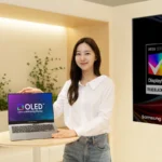 Samsung’s New Laptop OLED Hits 1,600 Nits and Lasts Twice as Long 3 A presenter displaying a Samsung OLED laptop on a wooden table, next to a large VESA DisplayHDR True Black 1400 certification poster.