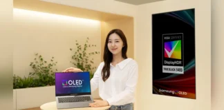 Samsung’s New Laptop OLED Hits 1,600 Nits and Lasts Twice as Long A presenter displaying a Samsung OLED laptop on a wooden table, next to a large VESA DisplayHDR True Black 1400 certification poster.
