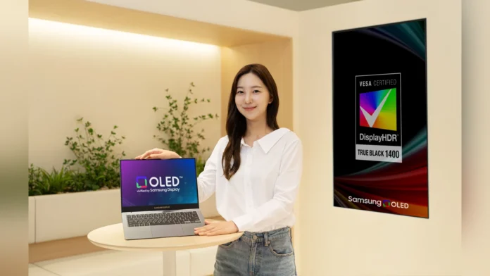 A presenter displaying a Samsung OLED laptop on a wooden table, next to a large VESA DisplayHDR True Black 1400 certification poster.